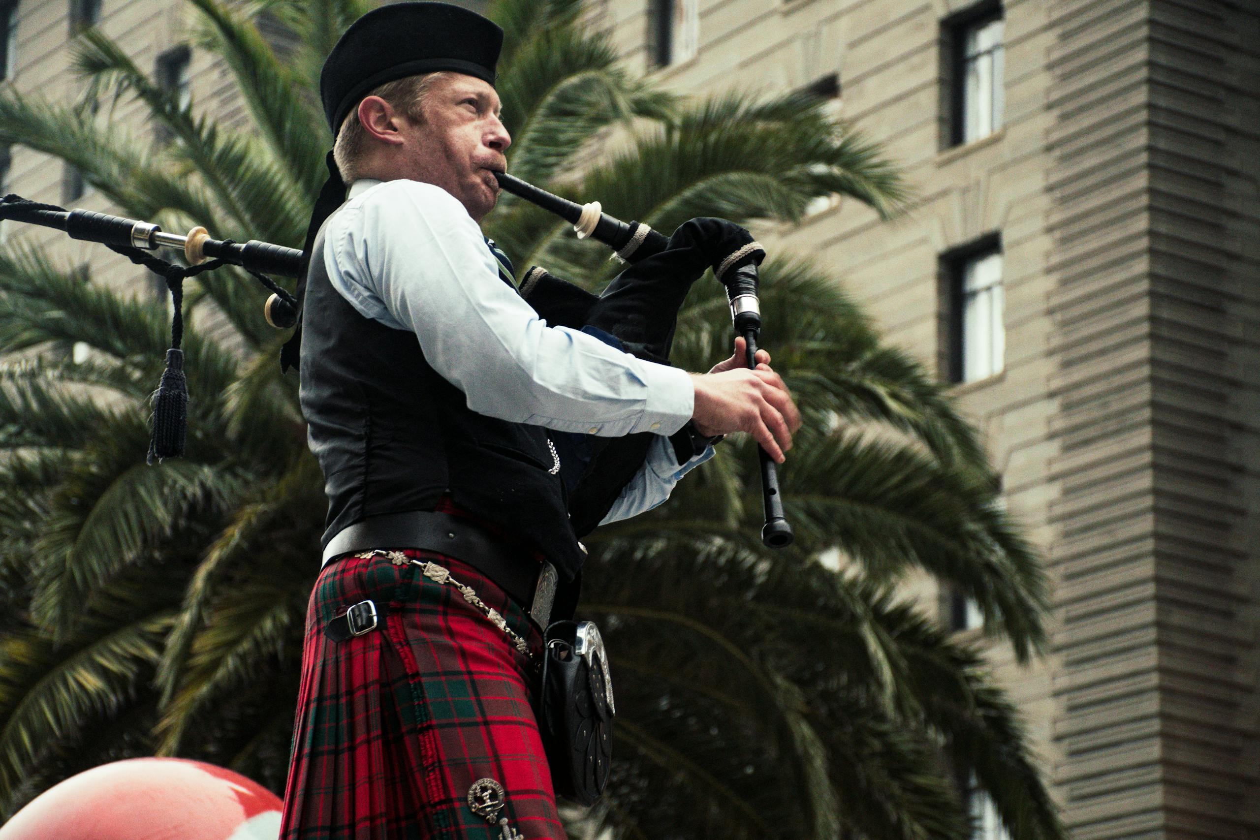 A male bagpiper playing outdoors, wearing traditional Scottish attire.